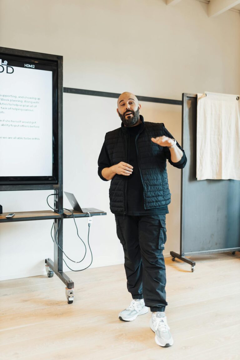 Bald man presenting in office with screen showing data, wearing black attire.